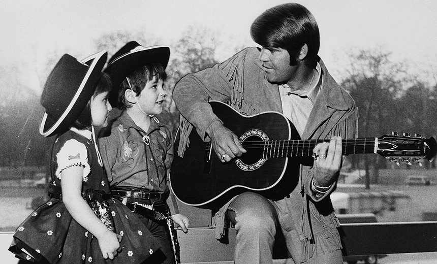 Country singer Glen Campbell playing the guitar for two young fans, Renata Romoli and Francis Bradford, wearing cowboys hats, outside the Hilton Hotel, April 23rd 1970. (Photo by Central Press/Hulton Archive/Getty Images)