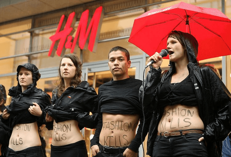 young white people wearing black standing in front of an H& M store with their black shirts hitched up to show FASHION KILLS written across their tummies