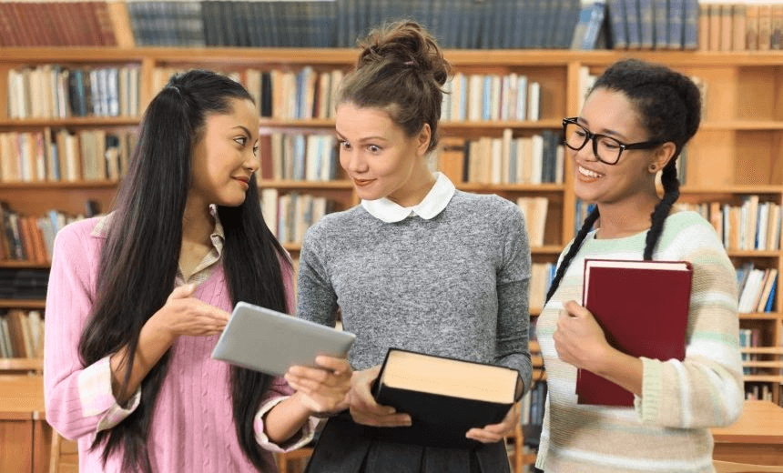 Three girls standing at library. All wear casual clothes. One with hair in ponytails holding digital tablet, other with braids and eyeglasses. Bookshelves and desks with chairs, windows as background. 
