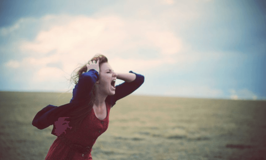 A young woman in a dress and sweater screaming in an empty field, on a cloudy afternon. 
