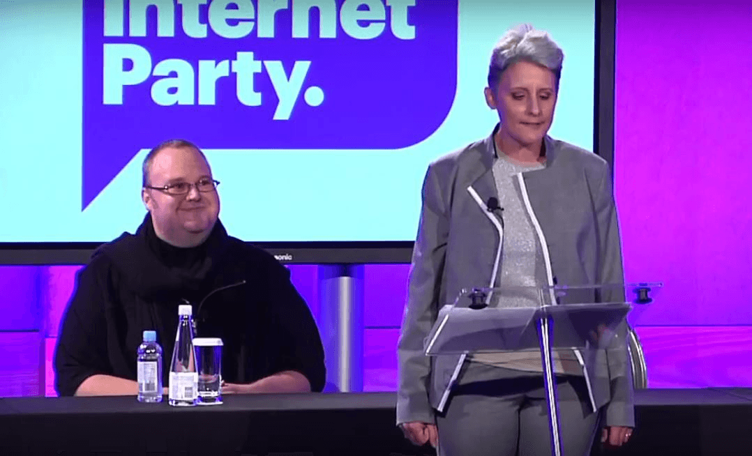 dotcom, in a black shirt, sitts behind laila harre, in a grey suit, with grey hair, speaking with an 'internet party' background