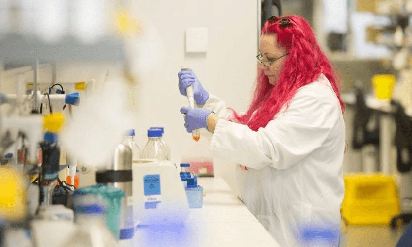 a pink haired white woman wearing a lab coat and gloves, putting something in a bottle with a pippette