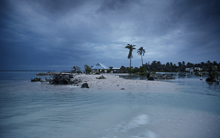 a stormy purple sky, with some houses on a pale islane and a pale turquoise swish of water in the foreground