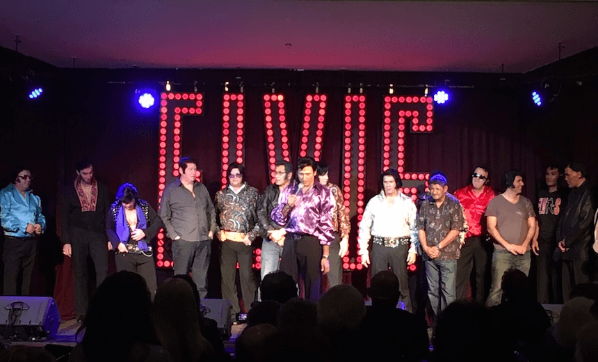 Photo of a line-up of Elvis impersonators on stage, huge neon ELVIS behind them
