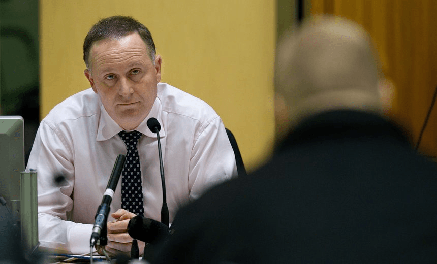 the back of a man in blurry foreground, with john key, a man with a white shirt and black tie, in the background