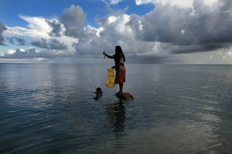 low, curly clouds linger over the flossy surface of the sea, where a figure with their back to the camera stands in ankle depth sea, holding a yellow bag