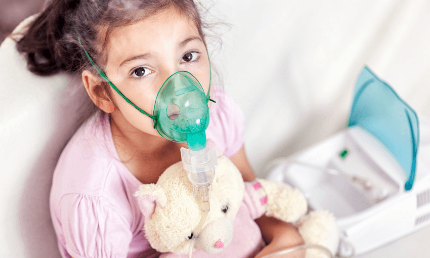 a child wearing a mask and clutching a teddy bear looking slightly worriedly towards the camera