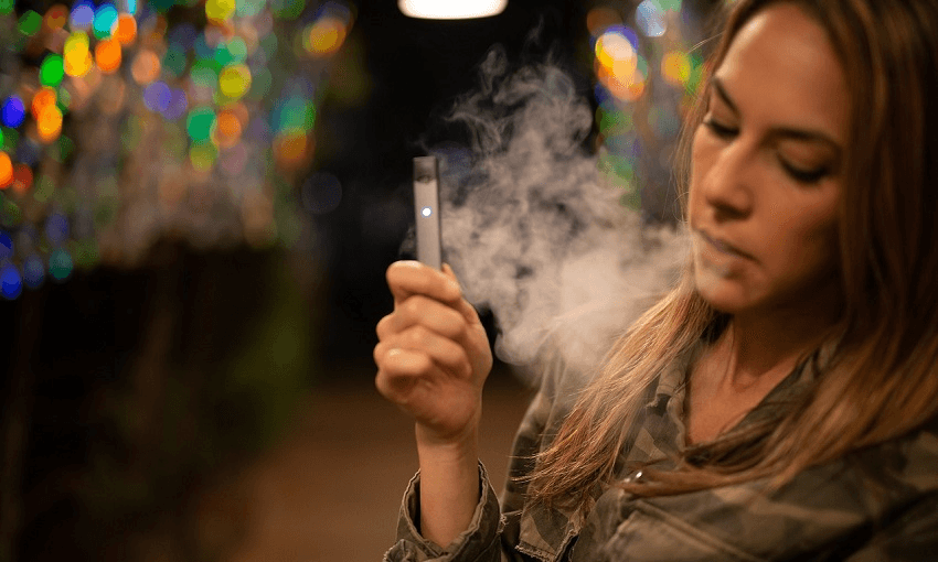 a young ish woman usning a vape. blonde, a cloud of smoke, and a dark background