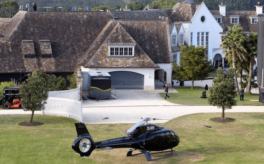 a helicopter landing on the lawn of a big house with manicured trees and a mottled green facade