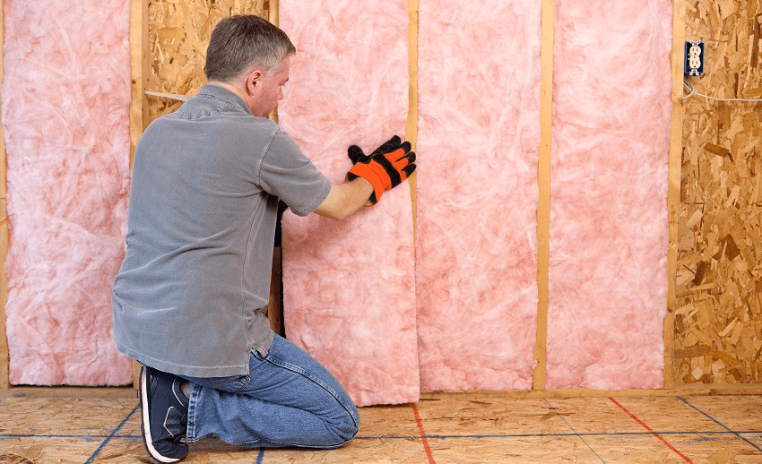 a man with a grey outfit and gloves eases a block of pink fibreglass into a wall recess