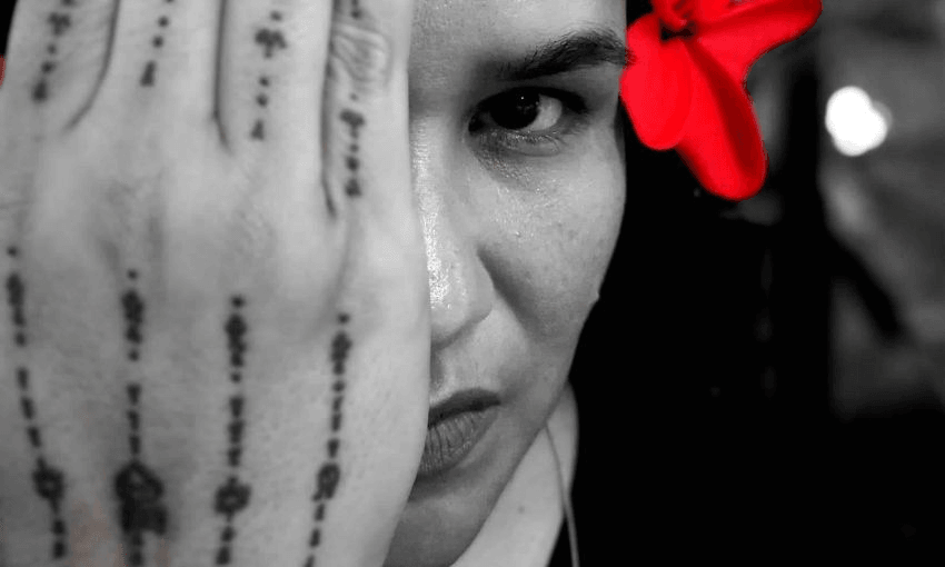 Photograph of a Samoan woman, hibiscus behind her ear, one hand over most of her face. Beautiful tatou on her hand.