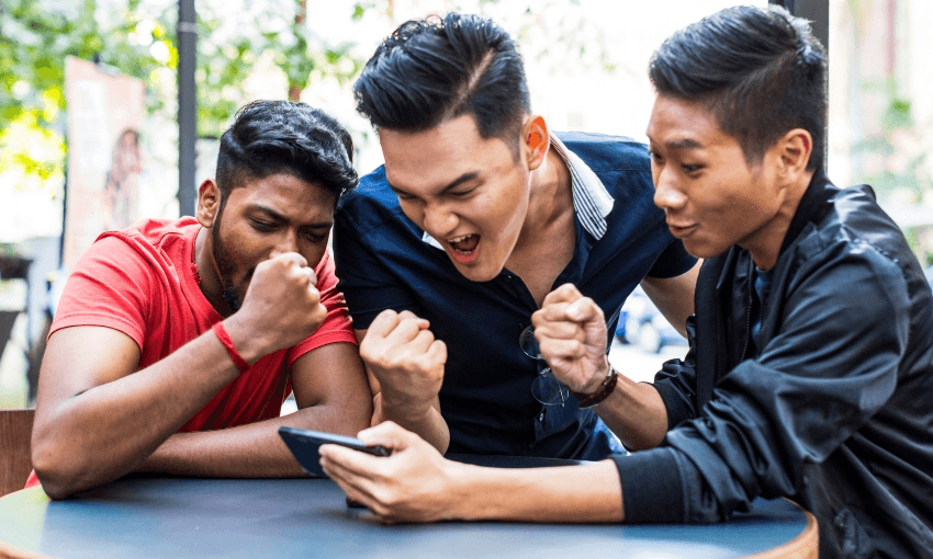 three young man, one looks indian and two look east asian, surrounding a phone and pumping their fists in victory