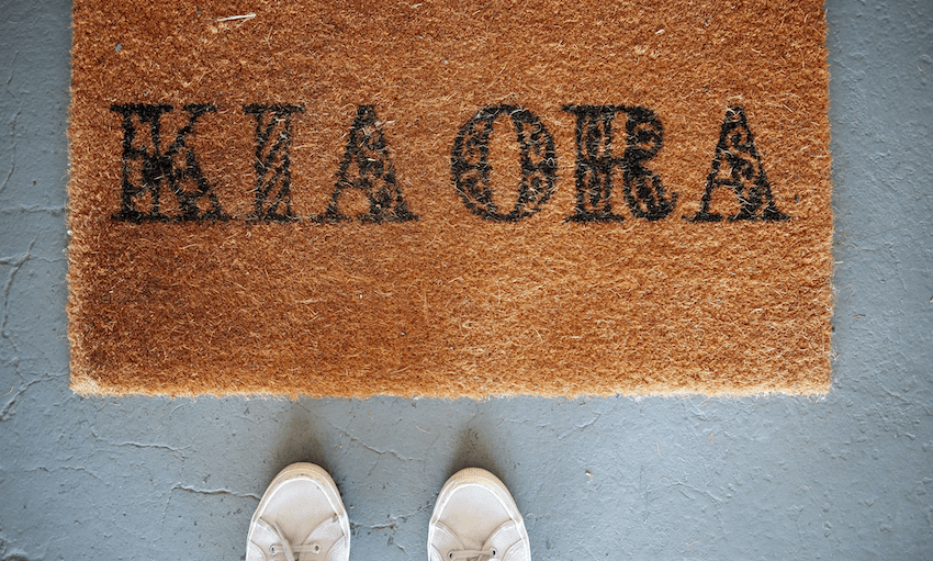 A brown doormat with the words "KIA ORA" in black letters, seen from above with a pair of white shoes visible at the bottom of the image.