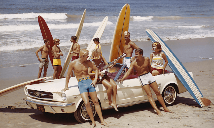 A group of surfers on a beach with a Ford Mustang car. 
