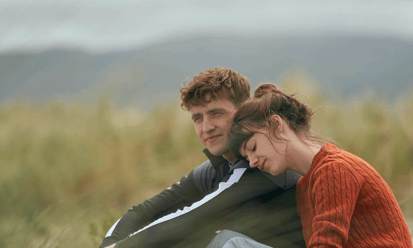 A grey day, tussocky field, a boy and a girl sit close, she resting her head on his shoulder. Both pale and beautiful.