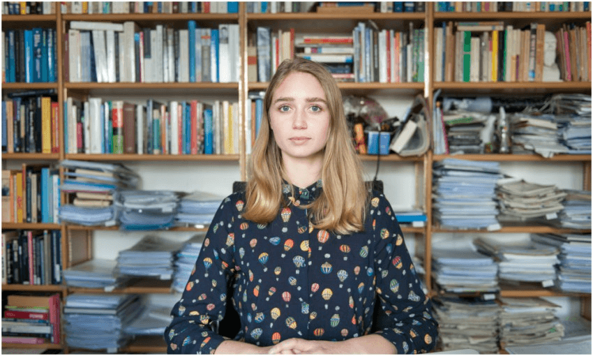 a young white woman looking nonplussed with bookshelves behind her