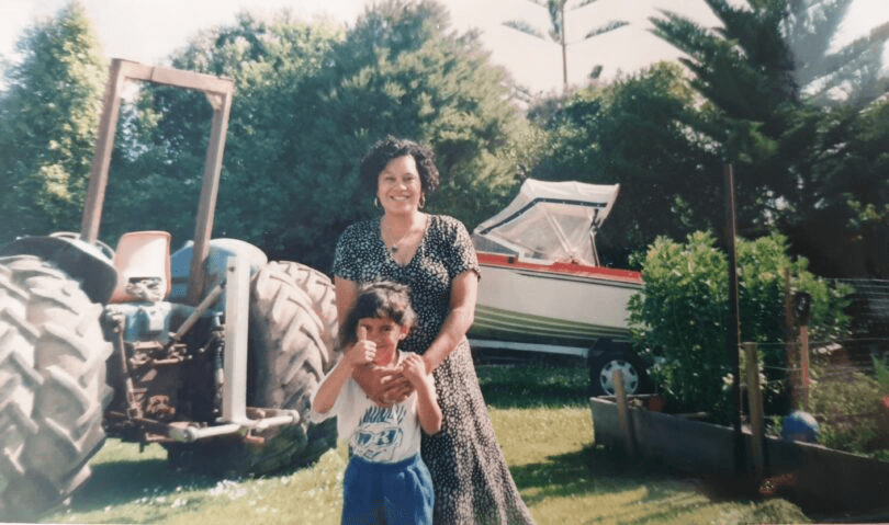 A woman and her young daughter smile at the camera. Backdrop is a garden with a tractor and boat, and broad beans.