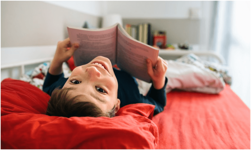 Boy lies on bed holding book and smiles upside-down at camera