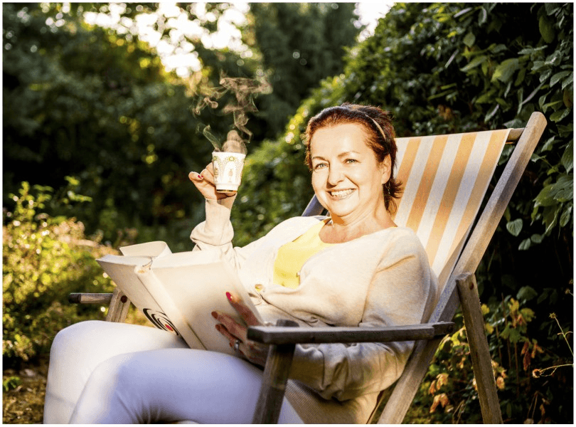 Middle-aged woman in deckchair with cup of tea and book, smiling. 