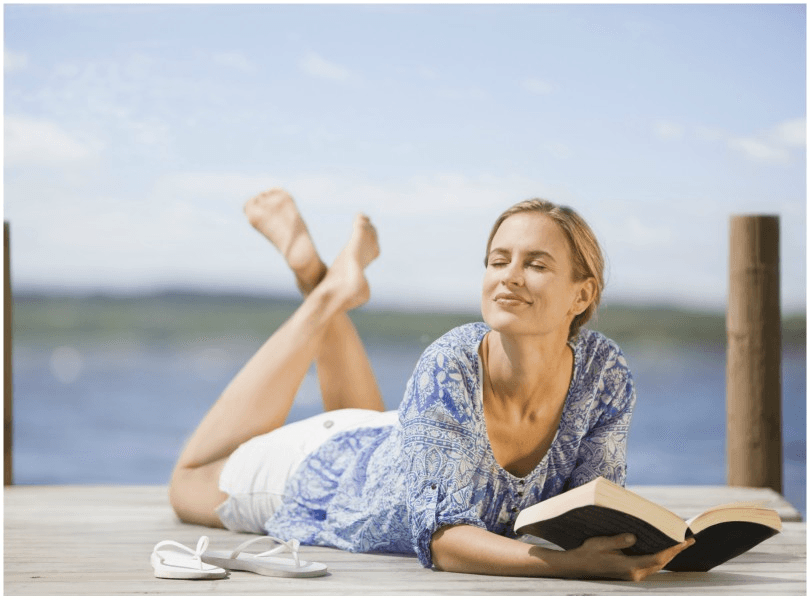 Woman lies tummy-down on a jetty, legs kicked up, propped on elbows with book. 