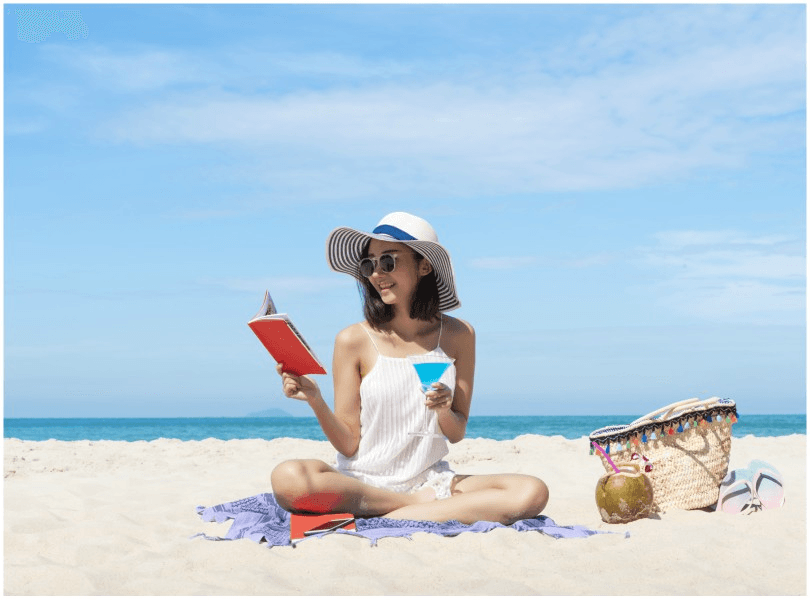Woman sitting cross-legged on beach, holding book in one hand and drink in the other.