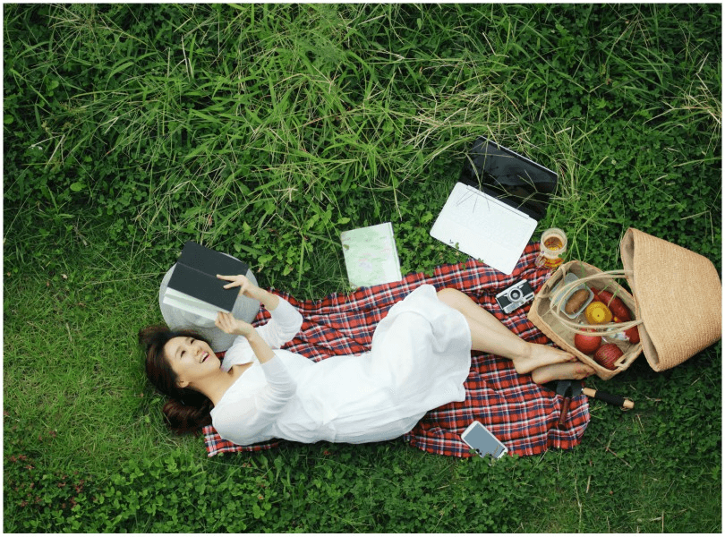 Woman lying on her back on picnic rug, curled up, holding book over her head.