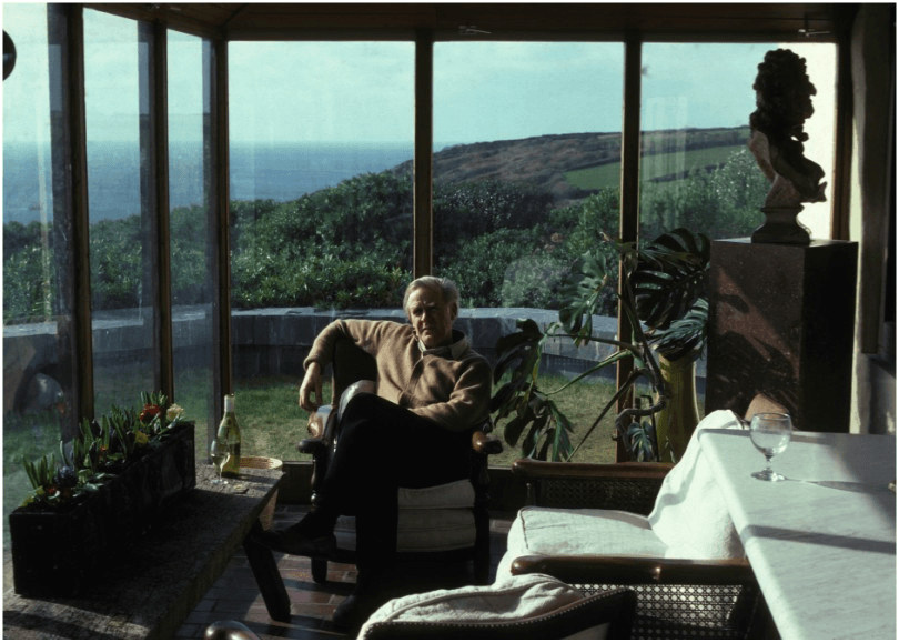 Man relaxes in conservatory with view over cliffs and sea. 
