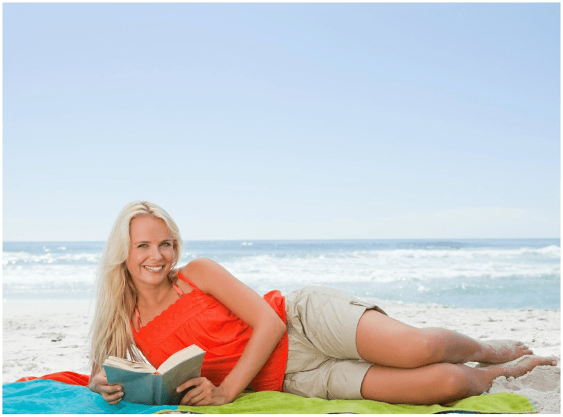 Woman lying on her side on the beach, smiling, reading.