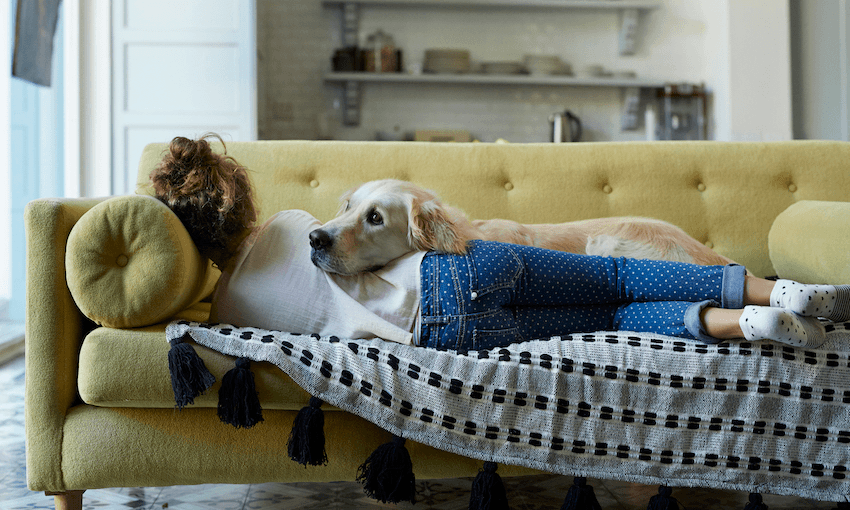 A young woman lies on a couch with her back to the camera, while a cute Golden retriever dog lies over her.