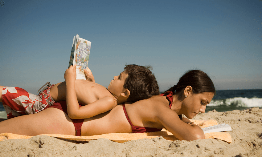 A woman lies on her front, reading, with a small boy lying on his back on top of her, reading. On a beach.