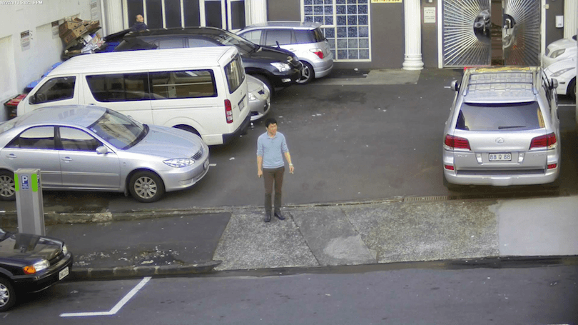 A lone man stands in a carpark, smoking, surrounded by cars. 