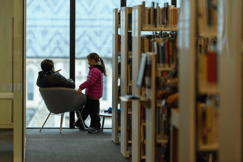 A child stands next to an adult in a chair, in a library