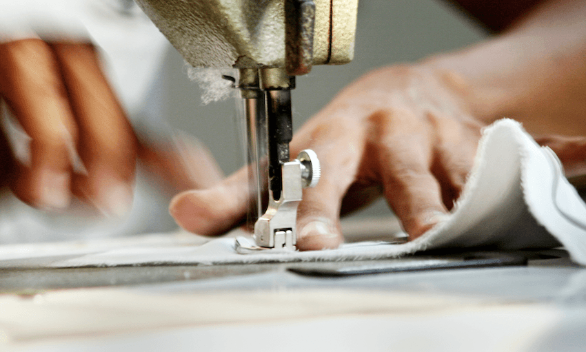 a foot of a sewing machine andsomeone's hands holding the edges of the seam steady as they stitch the fabric together