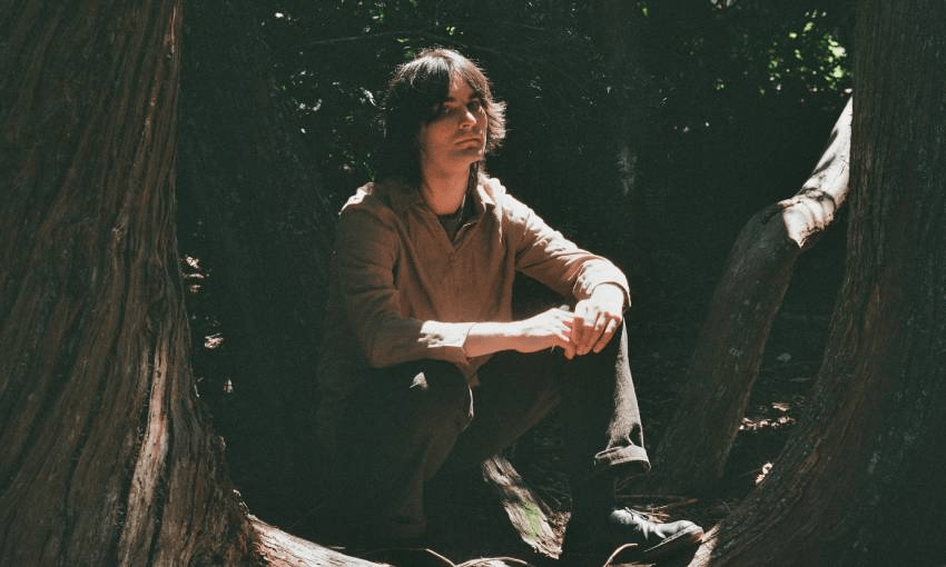 Kane Strang sits on a log in the woods, sunlight landing on his face and hands