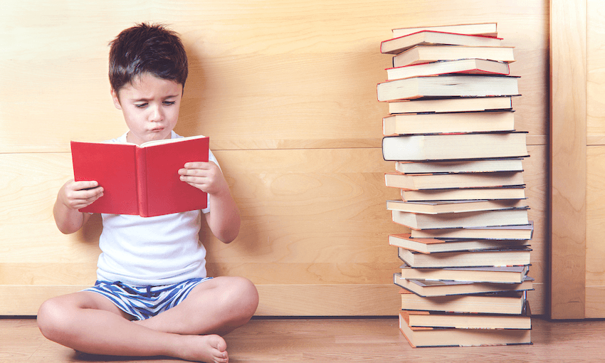 Cross-legged boy reads a book with a stack of books beside him.