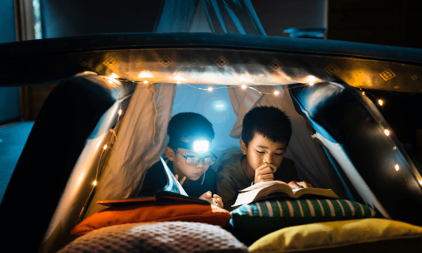 A cushion fort set up in a dark room, with fairy lights, two little boys reading books inside.