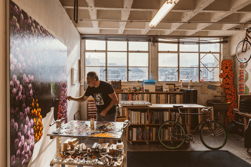 We're in an artist's studio, looking towards windows. The artist is leaning in to a huge canvas on the left-hand wall. Books and painting supplies everywhere. 