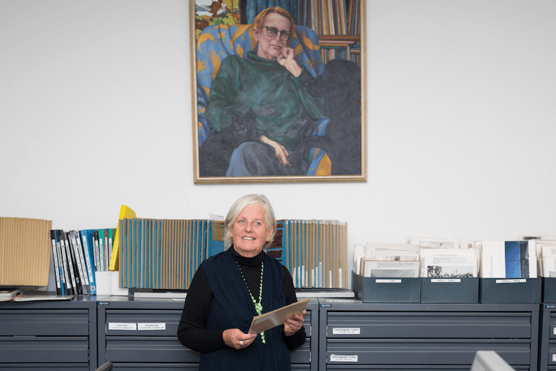 Senior woman stands in front of filing cabinets at a library, a huge painting hanging behind her.