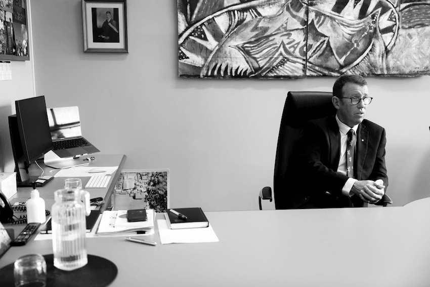 Patrick Drumm sits behind a large desk, eyes looking off camera, and talking to someone