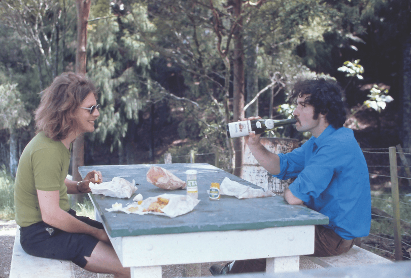 Two men at a picnic table, one swigging from a bottle, long hair and stubby shorts. 