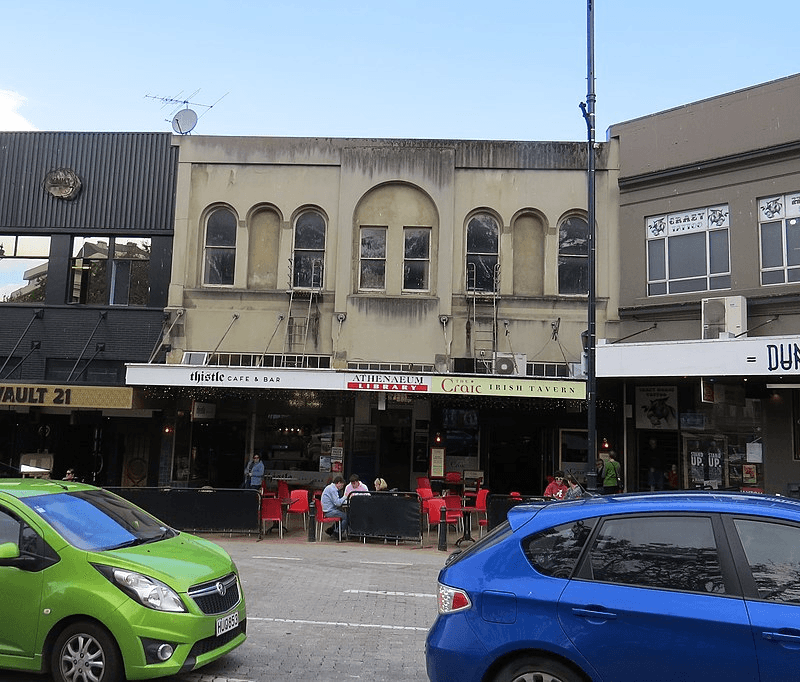 Street view, cars in foreground, then a road, cafe tables, and a row of old buildings at rear. 