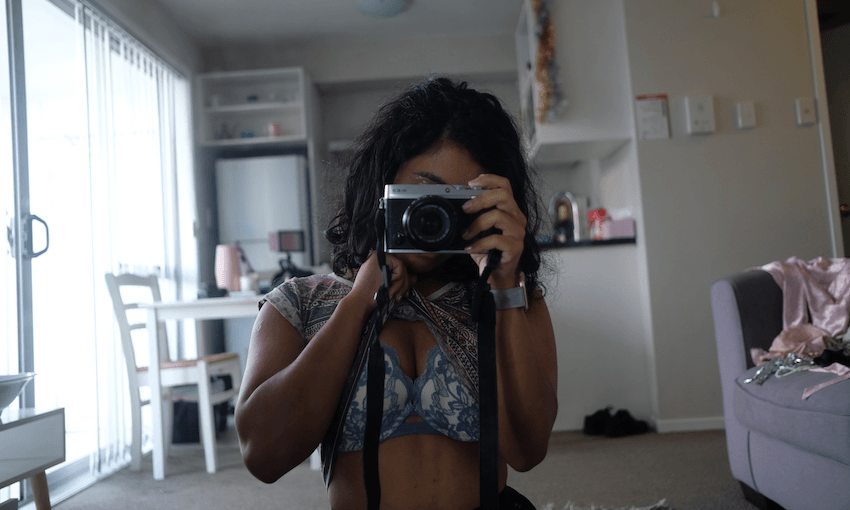 A woman stands in an apartment, wearing only a bra, face obscured by a camera.