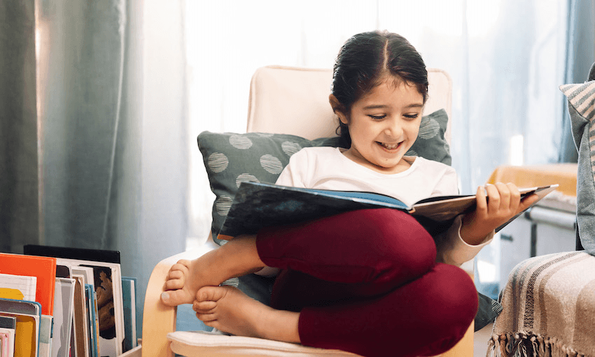 Photograph of a young girl curled up in a chair, barefoot, beaming as she reads a picture book.