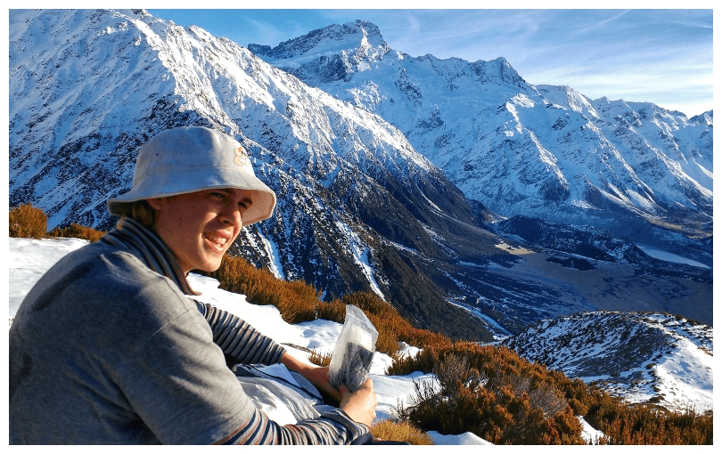 A young man sits on a snowy ridge, looking out over bare slopes and snow. 