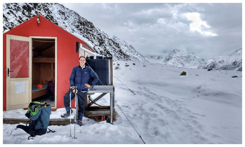 A young man in the snow, standing beside a bright red hut. 