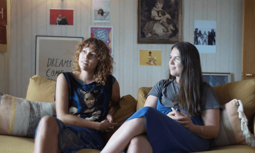 Nicole Gaffney and Billie Rogers sit side-by-side on a yellow couch, looking off-camera to the left.