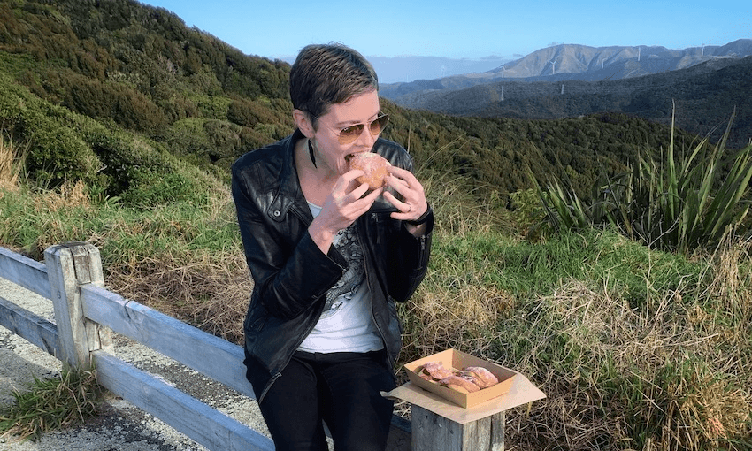 Woman sits on wooden fence biting into huge donut, box of donuts in front of her. Beautiful hills and sky in background.
