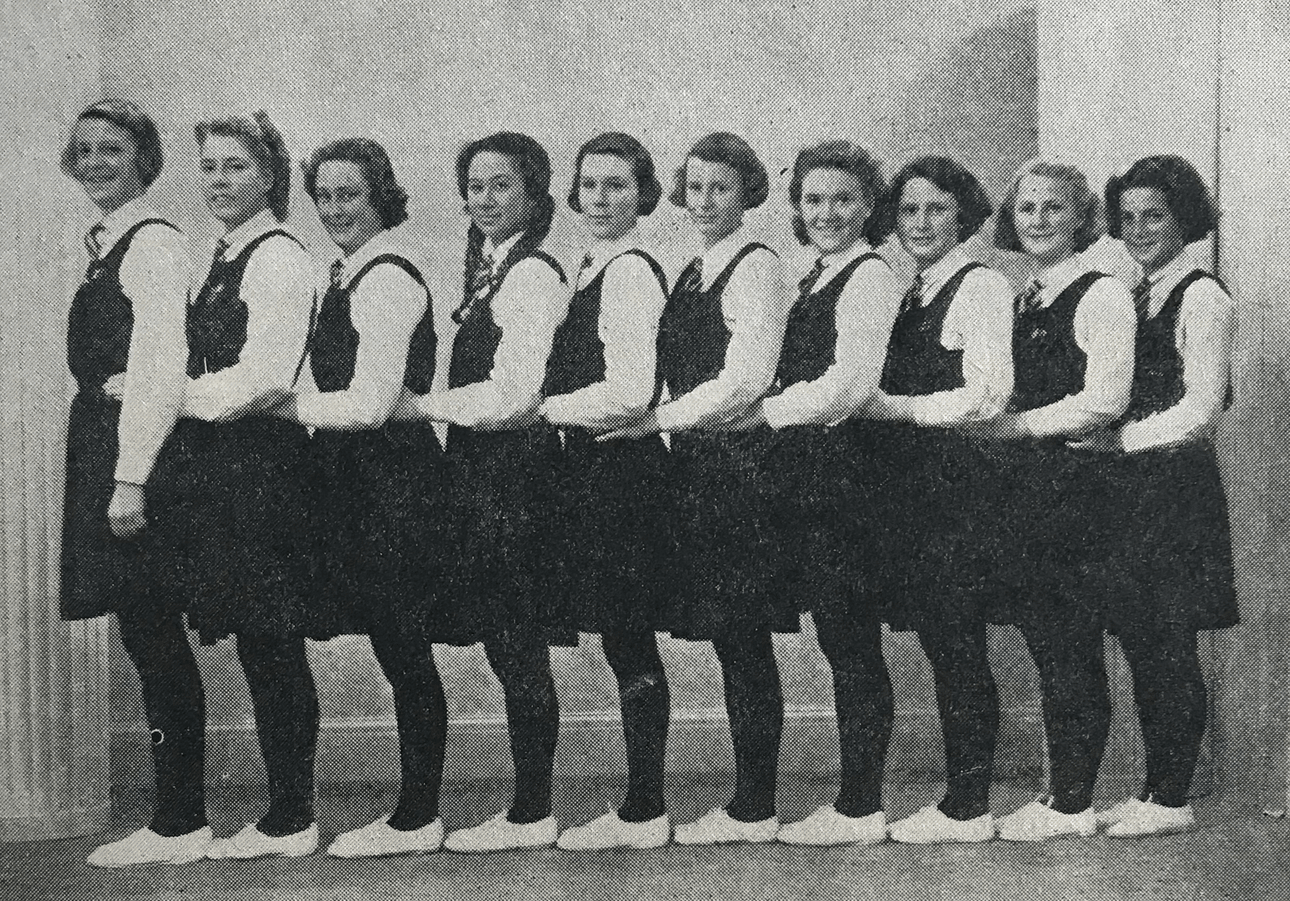 Black and white photograph of 10 schoolgirls in uniform, standing in a line with hands on the hips of the girl in front. One Māori girl, fourth from left.