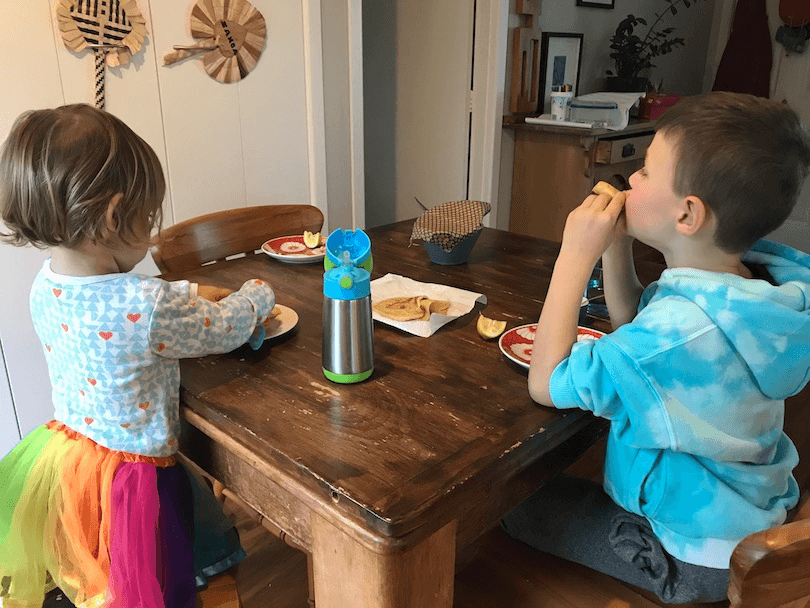 A kitchen table, two kids happily eating pancakes