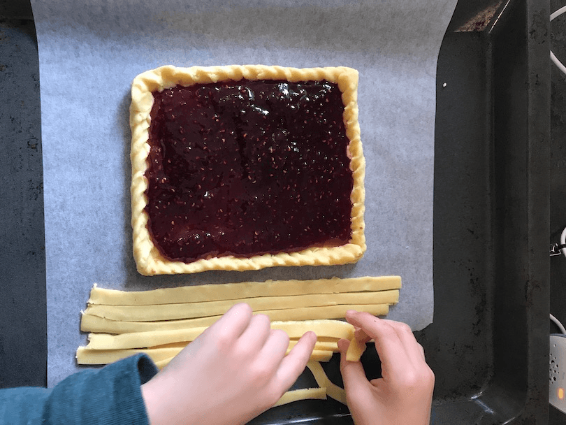 A child's hands pick up strips of pastry ready to arrange on a jam-topped tart